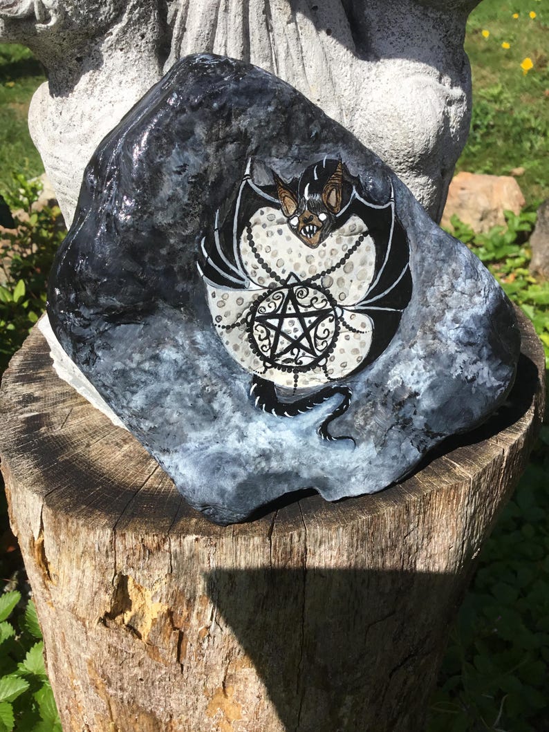 May include: A hand-painted rock featuring a black bat with white accents and a pentagram design. The bat is centered on a white and gray background, with a chain detail. The rock is dark gray and black.
