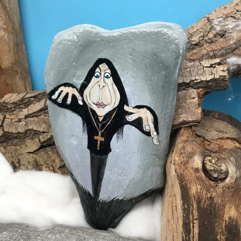 May include: A hand-painted rock depicts a cartoon figure in a black robe and gold cross necklace. The figure has exaggerated features and is painted on a grey, irregularly shaped rock. The background includes wood and a blue backdrop.