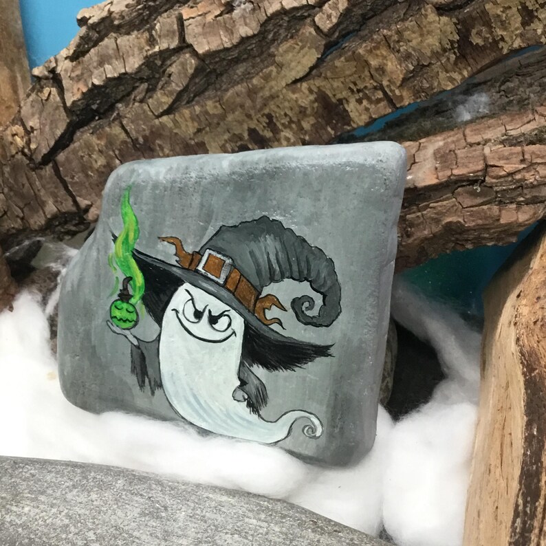 Peut inclure: Pierre grise peinte &agrave; la main repr&eacute;sentant un fant&ocirc;me de dessin anim&eacute; portant un chapeau de sorci&egrave;re avec une bande et une boucle marron. Le fant&ocirc;me tient une bouteille de potion verte avec de la fum&eacute;e verte. L'&oelig;uvre est plac&eacute;e sur un fond de bois et de coton.