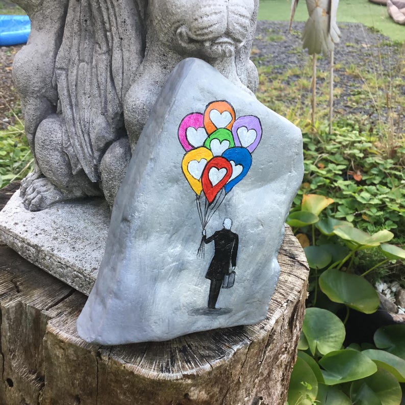 May include: A hand-painted rock features a person holding colorful heart-shaped balloons. The balloons are in shades of pink, orange, yellow, green, blue, and red. The person is depicted in black, wearing a coat and carrying a briefcase. The rock is gray.