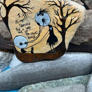 Peut inclure: Galet peint &agrave; la main avec une sc&egrave;ne fantaisiste. Deux personnages ronds aux yeux bleus sont reli&eacute;s par des lignes &agrave; une cr&eacute;ature noire ressemblant &agrave; un oiseau. Le texte "I always searched for a way out of the cruel world" est &eacute;crit sur le galet. Le fond est beige avec des silhouettes d'arbres noirs.