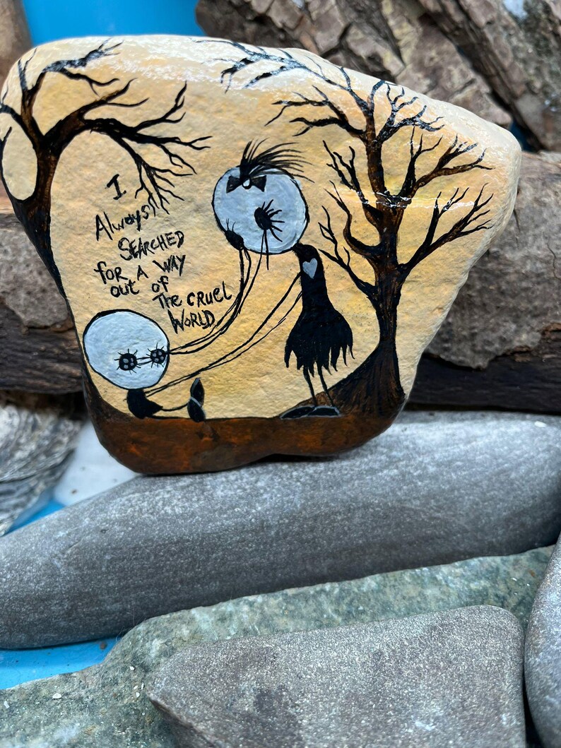 Peut inclure: Galet peint &agrave; la main avec une sc&egrave;ne fantaisiste. Deux personnages ronds aux yeux bleus sont reli&eacute;s par des lignes &agrave; une cr&eacute;ature noire ressemblant &agrave; un oiseau. Le texte "I always searched for a way out of the cruel world" est &eacute;crit sur le galet. Le fond est beige avec des silhouettes d'arbres noirs.