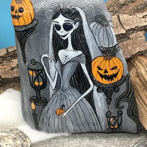 K&ouml;nnte beinhalten: Handbemalte Felskunst mit einer gotischen Braut mit schwarzen Haaren, Schleier und grauem Kleid. Das Kunstwerk zeigt K&uuml;rbisse, eine Kerze und eine Laterne. Die Farbpalette ist haupts&auml;chlich grau, orange und schwarz und erzeugt ein gruseliges Halloween-Thema.