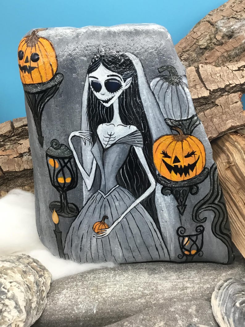 K&ouml;nnte beinhalten: Handbemalte Felskunst mit einer gotischen Braut mit schwarzen Haaren, Schleier und grauem Kleid. Das Kunstwerk zeigt K&uuml;rbisse, eine Kerze und eine Laterne. Die Farbpalette ist haupts&auml;chlich grau, orange und schwarz und erzeugt ein gruseliges Halloween-Thema.