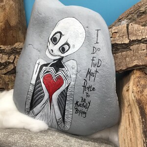 May include: A hand-painted gray rock features a skeletal figure holding a red heart. The figure has a white head and is dressed in black and white. The text reads, "I do find most people to be awfully boring."
