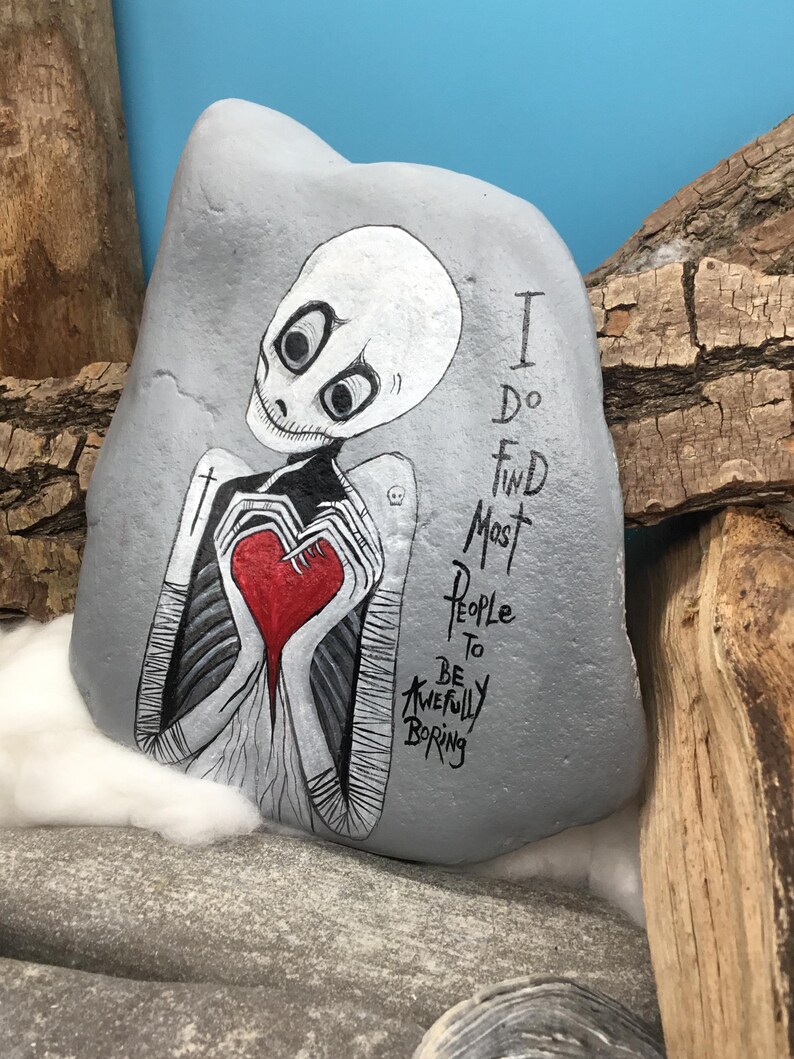 May include: A hand-painted gray rock features a skeletal figure holding a red heart. The figure has a white head and is dressed in black and white. The text reads, "I do find most people to be awfully boring."