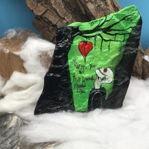 May include: Hand-painted rock art featuring a green and black design with a red heart and the words "Thank You for this Wonderful Painful Life." The artwork includes a skeleton figure and tree branches.