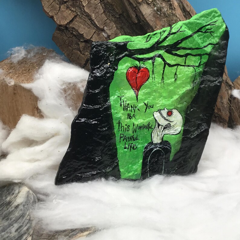May include: Hand-painted rock art featuring a green and black design with a red heart and the words "Thank You for this Wonderful Painful Life." The artwork includes a skeleton figure and tree branches.