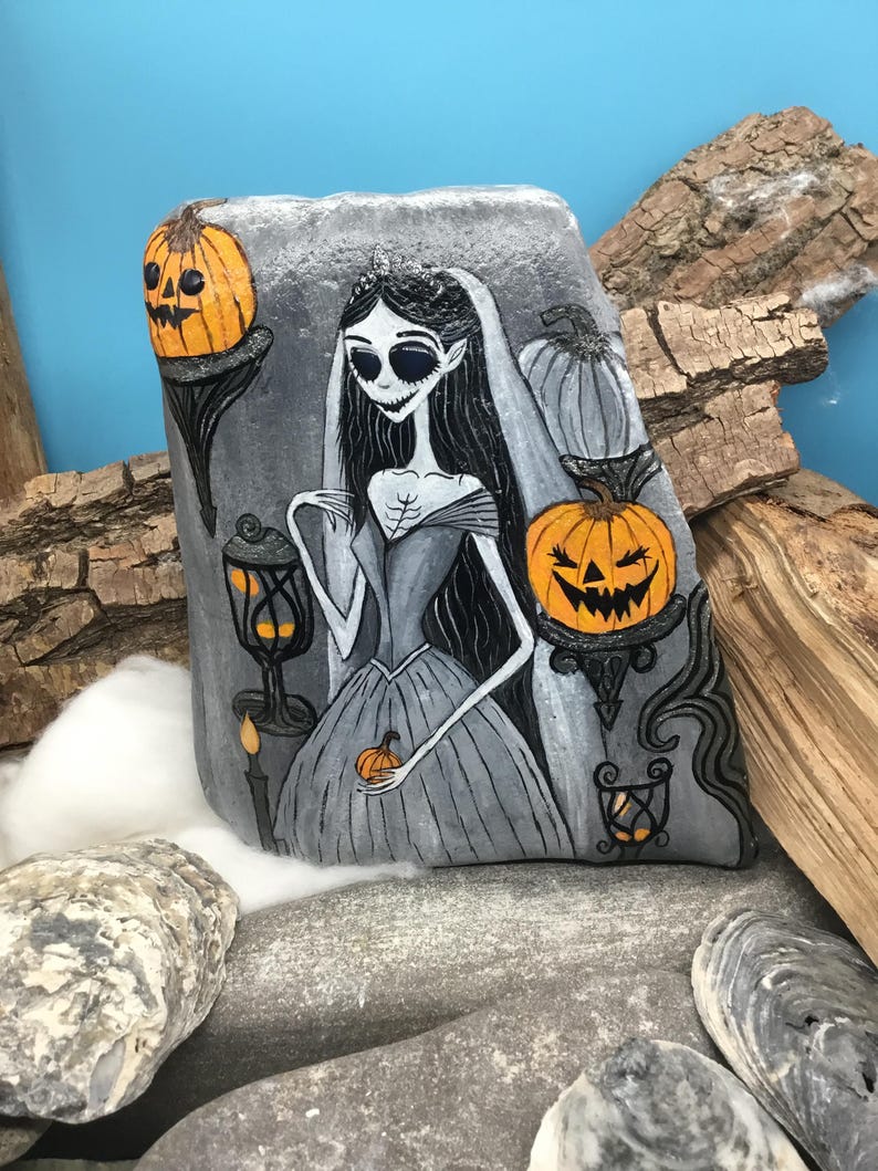 K&ouml;nnte beinhalten: Handbemalte Felskunst mit einer gotischen Braut mit langen schwarzen Haaren und einem grauen Kleid, die einen kleinen K&uuml;rbis h&auml;lt. Der Felsen ist mit K&uuml;rbislaternen und einer Kerze verziert, ideal f&uuml;r Halloween-Dekorationen.