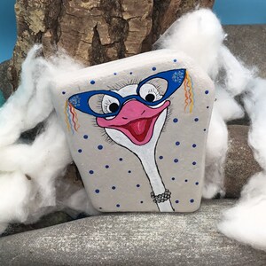 May include: Hand-painted rock art of an ostrich wearing blue glasses with a pink beak and a white neck. The rock is painted with a grey background and blue polka dots.