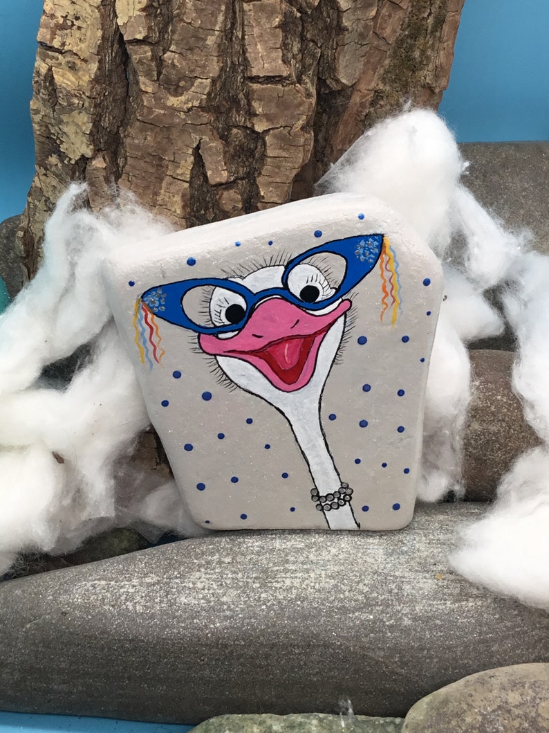 May include: Hand-painted rock art of an ostrich wearing blue glasses with a pink beak and a white neck. The rock is painted with a grey background and blue polka dots.