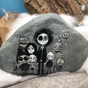 Puede incluir: Una piedra gris lisa con una ilustraci&oacute;n pintada a mano de un grupo de personajes estilizados y monocrom&aacute;ticos con ojos grandes y rostros expresivos. La obra incluye una figura central alta rodeada de varias criaturas m&aacute;s peque&ntilde;as.