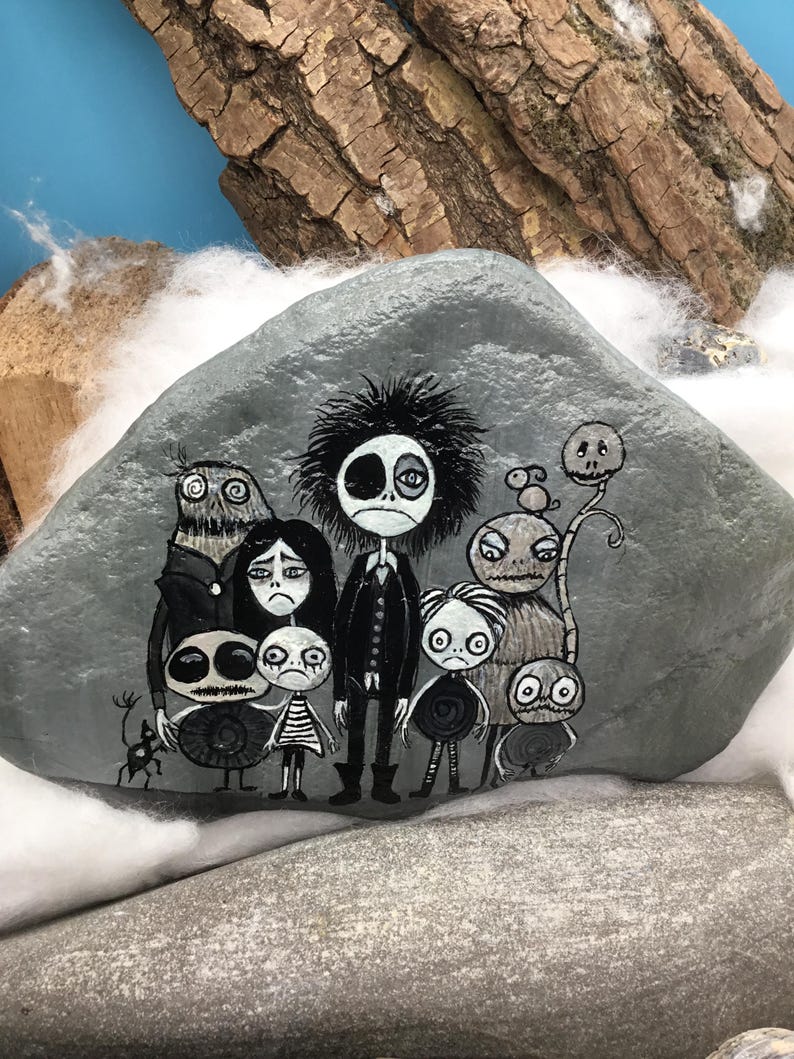 Puede incluir: Una piedra gris lisa con una ilustraci&oacute;n pintada a mano de un grupo de personajes estilizados y monocrom&aacute;ticos con ojos grandes y rostros expresivos. La obra incluye una figura central alta rodeada de varias criaturas m&aacute;s peque&ntilde;as.