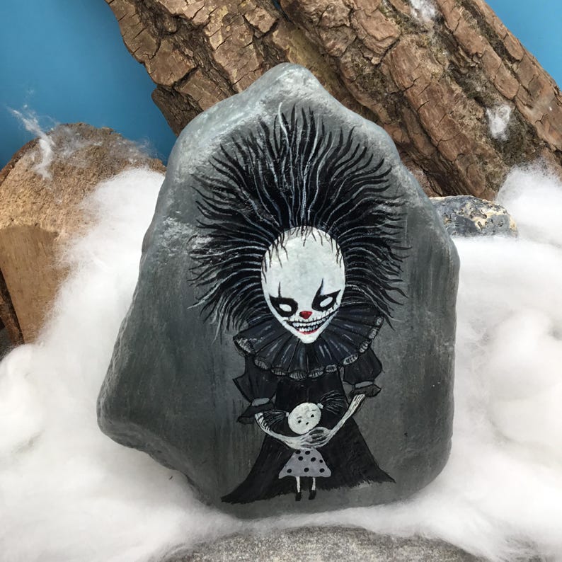 May include: A grey stone painted with a detailed illustration of a clown figure with spiky black hair, a ruffled collar, and a wide grin, holding a small doll with a polka dot dress. The artwork features high contrast black, white, and red paint.