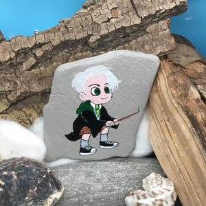May include: Hand-painted rock art featuring a cartoon character with white hair, green eyes, and a green and black robe. The character is holding a wand. The rock is gray and sits among other rocks, wood, and a seashell.