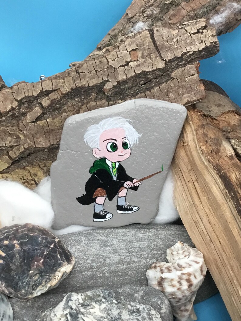 May include: Hand-painted rock art featuring a cartoon character with white hair, green eyes, and a green and black robe. The character is holding a wand. The rock is gray and sits among other rocks, wood, and a seashell.