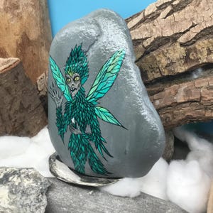 May include: A hand-painted, gray stone features a detailed illustration of a fairy with green and teal wings and body. The fairy has a stern expression. The stone is displayed on a shell, with wood and cotton in the background.