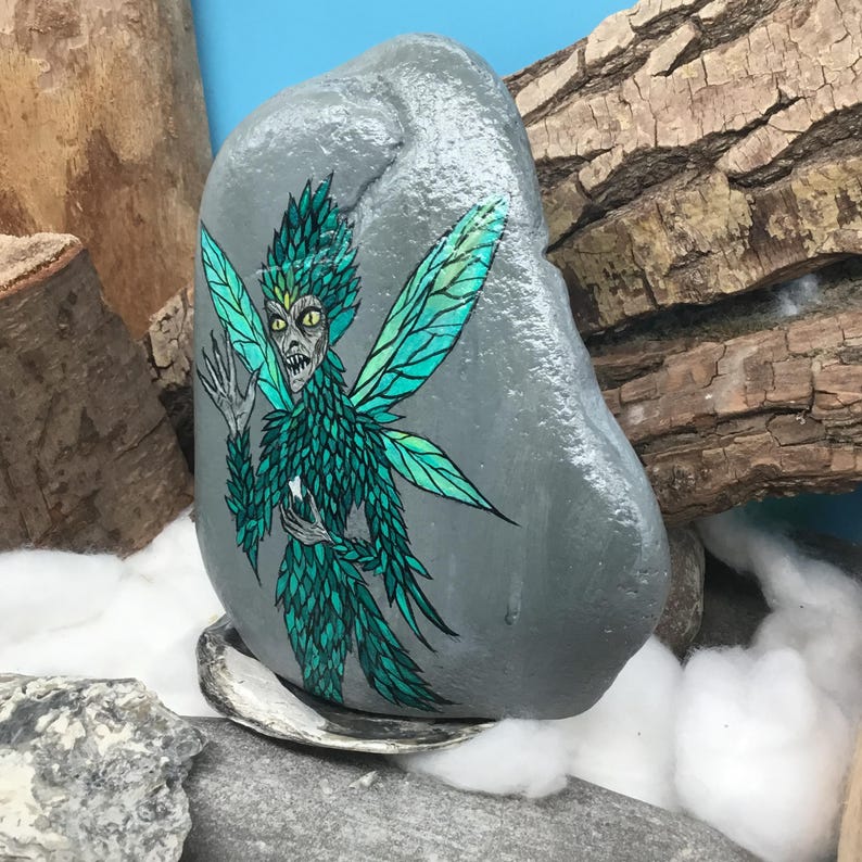 May include: A hand-painted, gray stone features a detailed illustration of a fairy with green and teal wings and body. The fairy has a stern expression. The stone is displayed on a shell, with wood and cotton in the background.