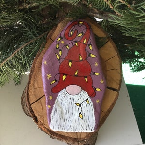 May include: Hand-painted rock art of a gnome wearing a red hat with yellow lights wrapped around it. The gnome has a white beard and a pink nose. The rock is painted purple with white stars.