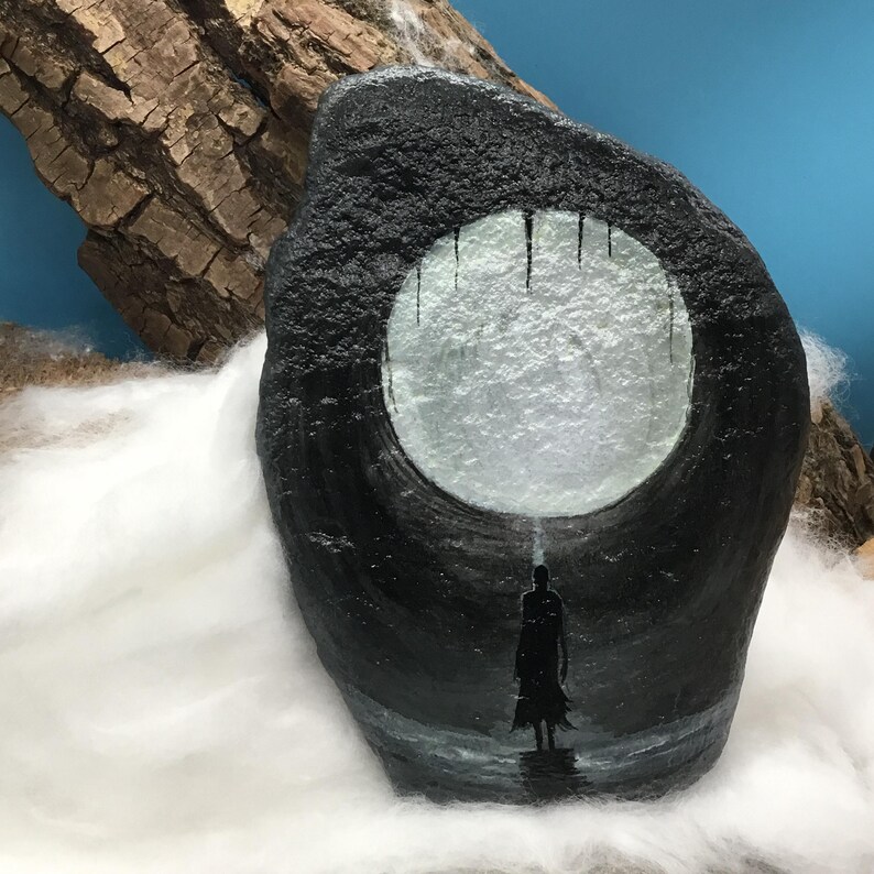 Pu&ograve; includere: Un'opera d'arte su roccia dipinta a mano in nero con una figura silhouette davanti a un'apertura circolare luminosa. L'opera d'arte ha una superficie strutturata ed &egrave; ambientata su uno sfondo bianco e blu.