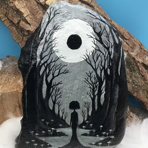 May include: Hand-painted rock art featuring a grayscale forest scene with a silhouetted figure. The artwork depicts a full moon and bare trees, creating a mystical and enchanting aesthetic. White flowers dot the foreground, adding detail to the dark landscape.