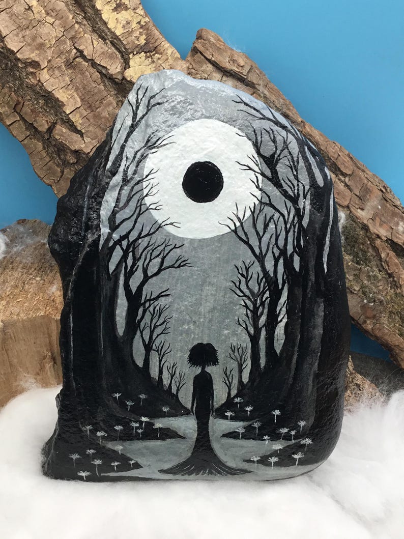 May include: Hand-painted rock art featuring a grayscale forest scene with a silhouetted figure. The artwork depicts a full moon and bare trees, creating a mystical and enchanting aesthetic. White flowers dot the foreground, adding detail to the dark landscape.