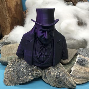 May include: A 3D-printed bust of a man in a top hat and formal wear, in a deep purple hue. The bust is displayed on rocks and oyster shells, with a backdrop of white fluffy material and a piece of wood.