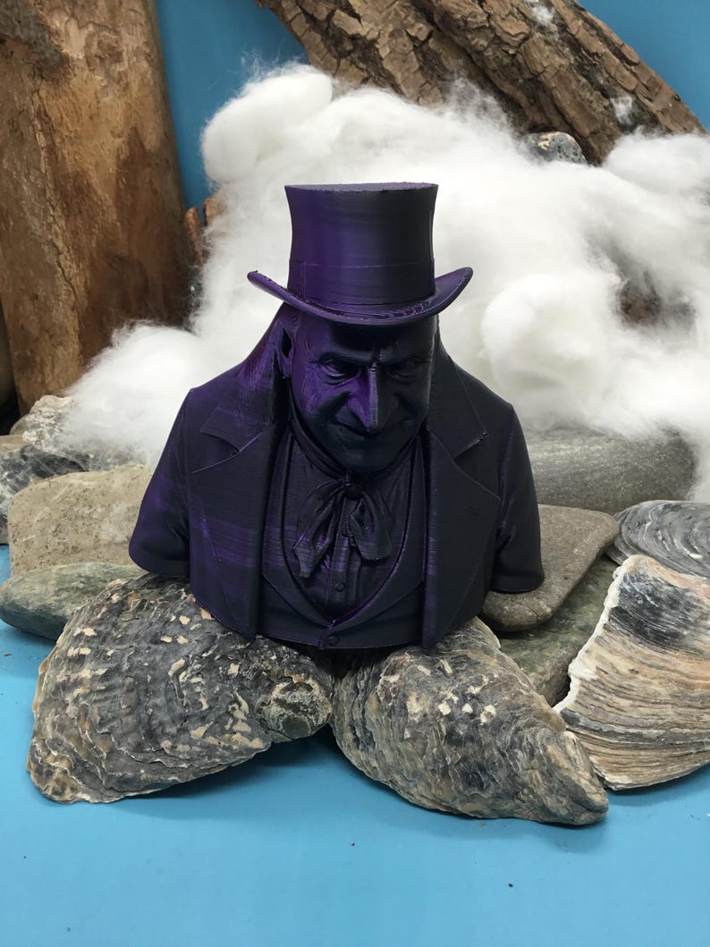 May include: A 3D-printed bust of a man in a top hat and formal wear, in a deep purple hue. The bust is displayed on rocks and oyster shells, with a backdrop of white fluffy material and a piece of wood.