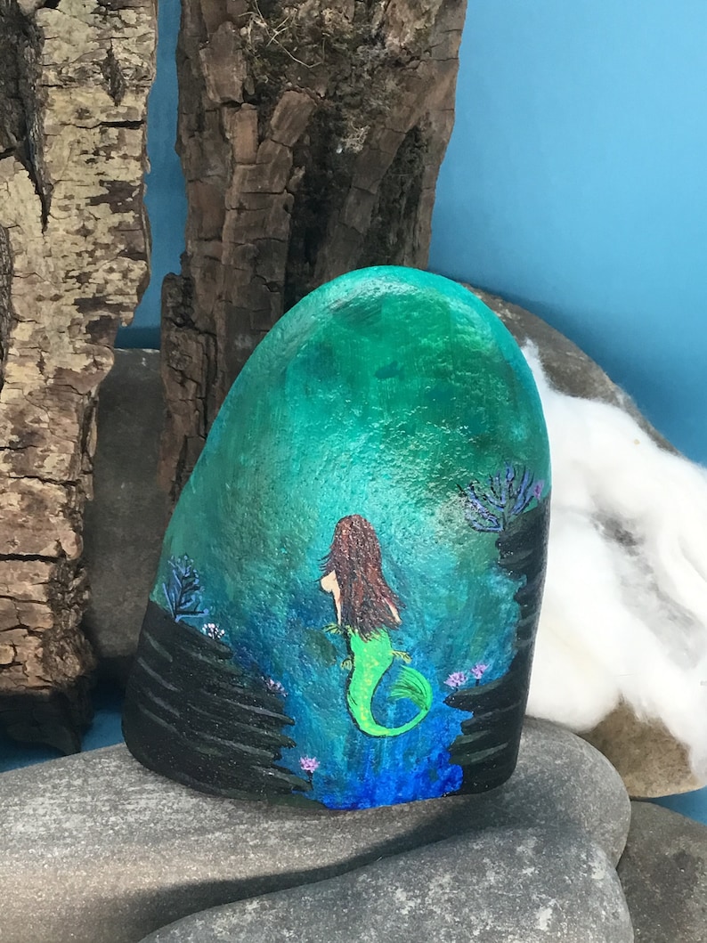 May include: Hand-painted rock art depicting a mermaid with a green tail and brown hair, swimming in a blue and green ocean scene with black rocks and purple coral.