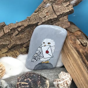 May include: Hand-painted grey stone featuring a white owl holding a letter with a red wax seal. The owl is perched on a grey rock, set against a blue background with seashells and driftwood. A unique, decorative item.