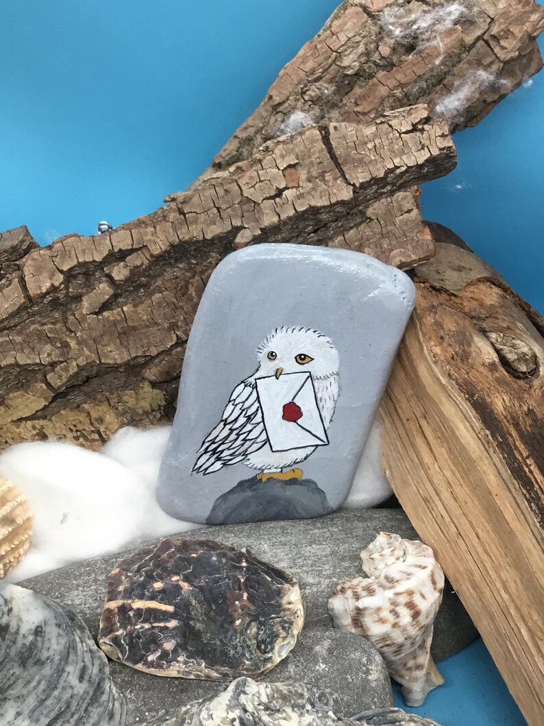 May include: Hand-painted grey stone featuring a white owl holding a letter with a red wax seal. The owl is perched on a grey rock, set against a blue background with seashells and driftwood. A unique, decorative item.