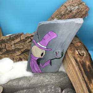 May include: A hand-painted, gray stone features a whimsical illustration of a skeleton figure in a purple top hat and coat, holding a crystal ball. The artwork is set against a blue backdrop with wood accents.