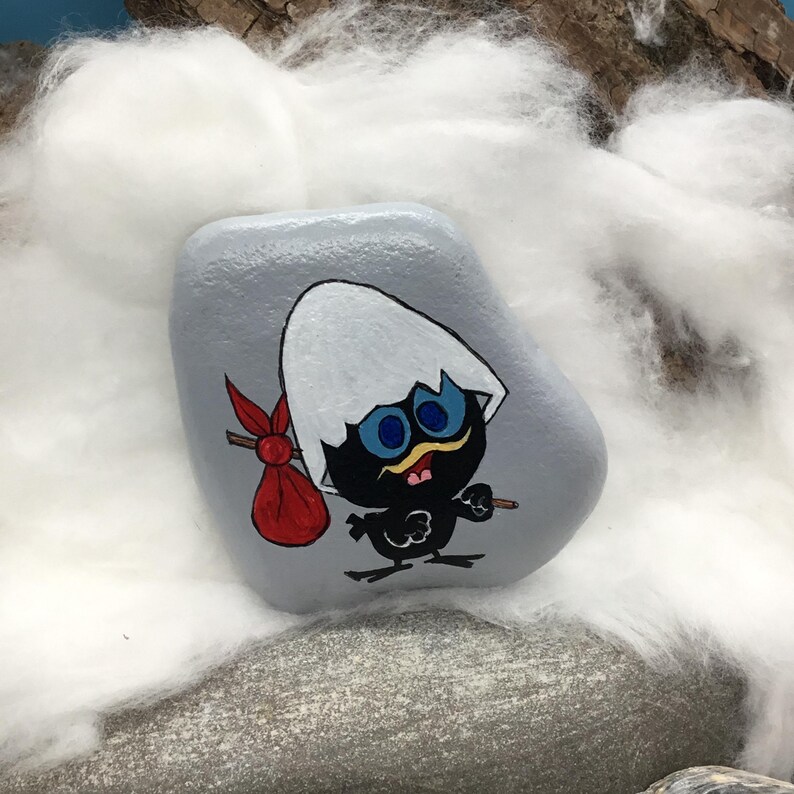 K&ouml;nnte beinhalten: Ein handbemalter, grauer Stein zeigt eine Cartoon-K&uuml;kenfigur mit einer zerbrochenen Eierschale auf dem Kopf. Das K&uuml;ken ist schwarz mit blauen Augen, einem gelben Schnabel und einem roten Sack. Das Kunstwerk ist vor einem Hintergrund aus wei&szlig;em Flaum und Steinen platziert.