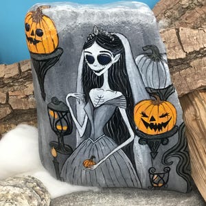 K&ouml;nnte beinhalten: Handbemalte Felskunst mit einer gruseligen Braut mit langen schwarzen Haaren, einer Tiara und einem grauen Kleid. Das Kunstwerk zeigt orangefarbene K&uuml;rbisse und eine Laterne, alles vor grauem Hintergrund. Eine einzigartige Halloween-Dekoration.