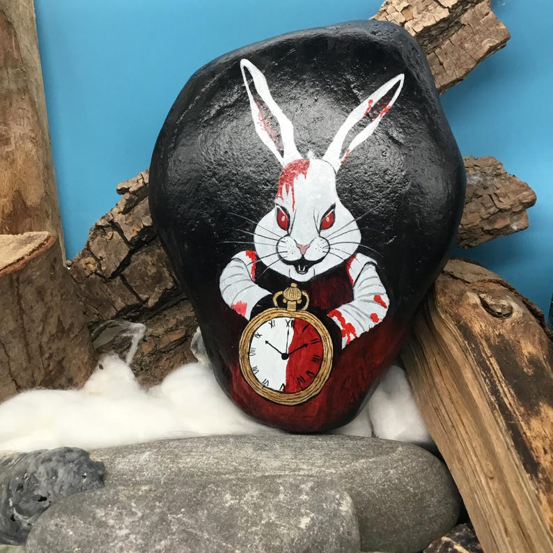 May include: A painted rock depicts a white rabbit with red eyes and blood-stained fur, clutching a pocket watch. The rabbit is set against a black backdrop. The rock is displayed with wood and other rocks.