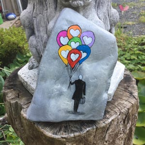 May include: Hand-painted gray rock art featuring a person in a black coat holding a bouquet of colorful heart-shaped balloons. The balloons are in shades of red, orange, yellow, green, blue, and purple. The rock is displayed on a tree stump.