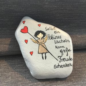 K&ouml;nnte beinhalten: Bemalter Stein mit einem Cartoon-Engel, der ein Herz an einem Stock h&auml;lt, umgeben von kleinen roten Herzen. Der Stein tr&auml;gt den deutschen Text "Selbst ein kleines L&auml;cheln kann gro&szlig;e Freude schenken."