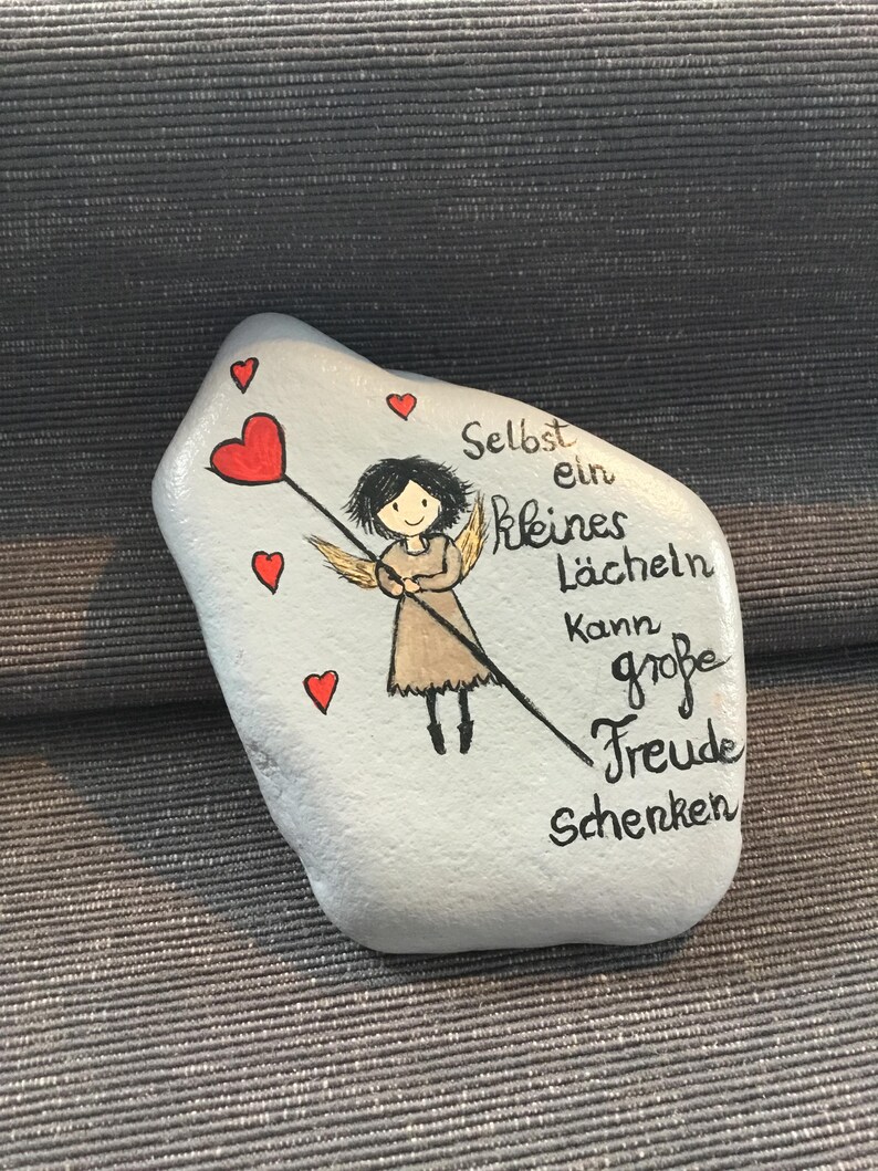 K&ouml;nnte beinhalten: Bemalter Stein mit einem Cartoon-Engel, der ein Herz an einem Stock h&auml;lt, umgeben von kleinen roten Herzen. Der Stein tr&auml;gt den deutschen Text "Selbst ein kleines L&auml;cheln kann gro&szlig;e Freude schenken."