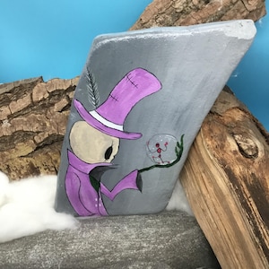 May include: Hand-painted gray stone featuring a skeleton figure in a purple top hat and coat, holding a crystal ball. The artwork includes a feather and a white brim on the hat. The background is a mix of wood and blue.