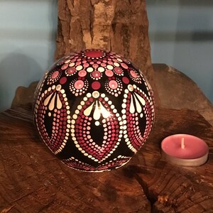 Op de afbeelding: Een zwarte bol met een roze en wit mandala erop geschilderd, die op een houten oppervlak staat met een roze kaars.