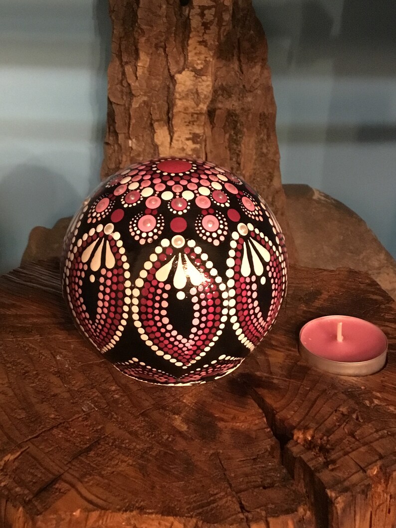 Op de afbeelding: Een zwarte bol met een roze en wit mandala erop geschilderd, die op een houten oppervlak staat met een roze kaars.