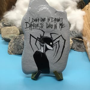 May include: A grey painted rock featuring a black ink illustration of a spindly, multi-limbed creature with a white eye. The text "I DON'T LIVE IN DARKNESS DARKNESS LIVES IN ME" is written in black ink above the creature on the surface of the stone.