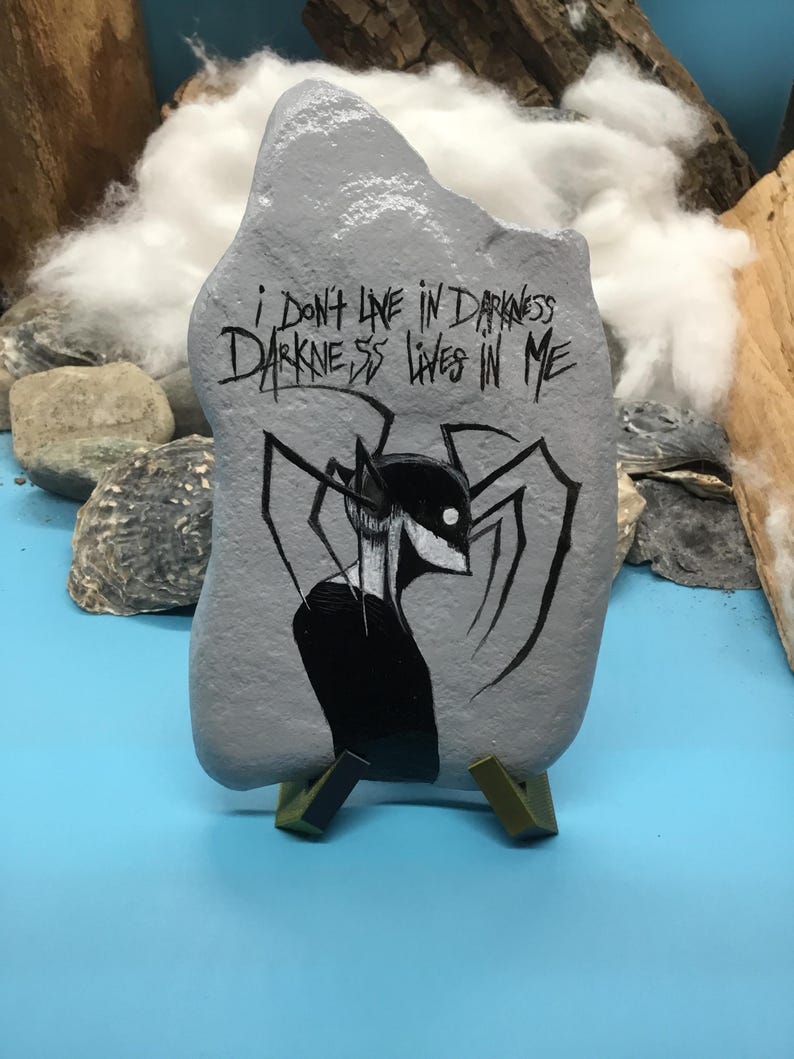 May include: A grey painted rock featuring a black ink illustration of a spindly, multi-limbed creature with a white eye. The text "I DON'T LIVE IN DARKNESS DARKNESS LIVES IN ME" is written in black ink above the creature on the surface of the stone.