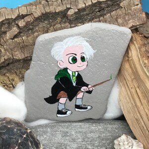 May include: Hand-painted rock art featuring a cartoon character with white hair, green eyes, and a green and black robe, holding a wand. The artwork is set against a backdrop of wood and shells, creating a whimsical, fantasy-themed piece.