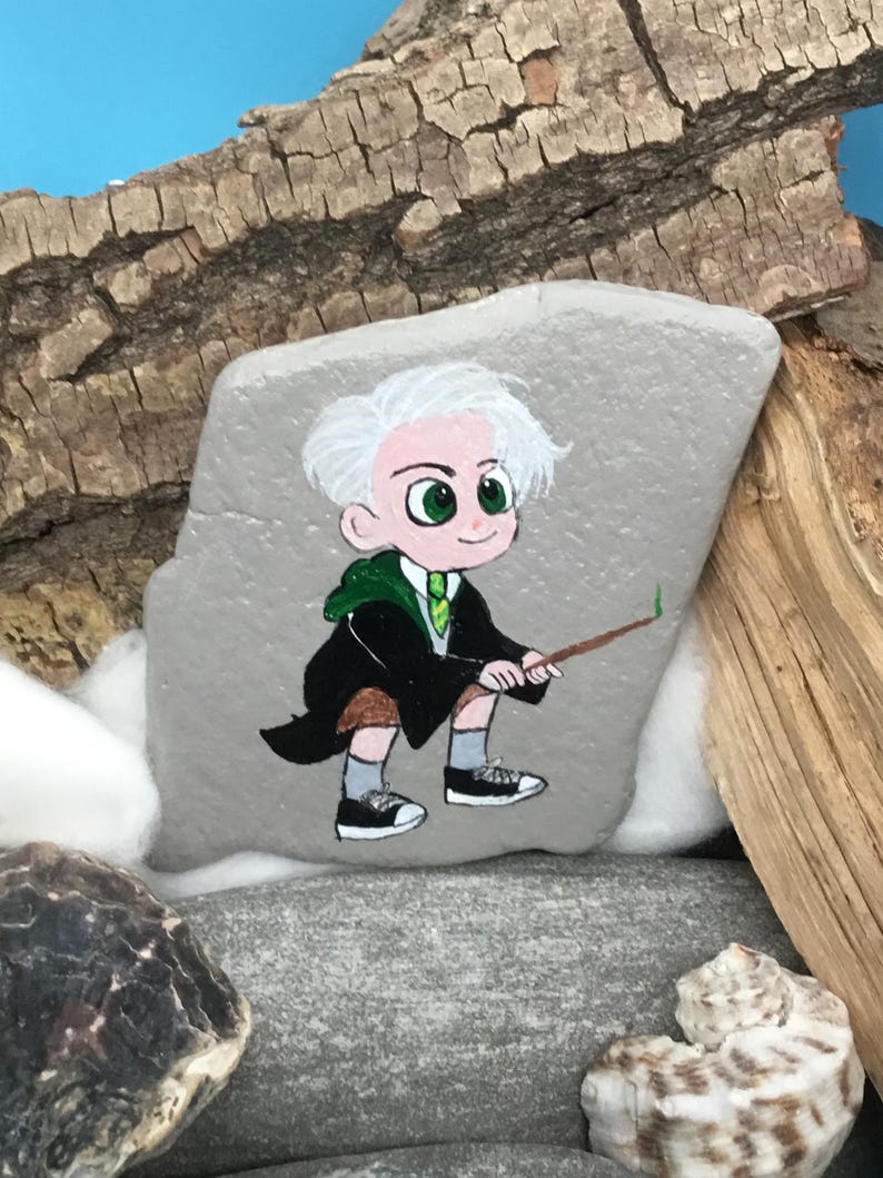 May include: Hand-painted rock art featuring a cartoon character with white hair, green eyes, and a green and black robe, holding a wand. The artwork is set against a backdrop of wood and shells, creating a whimsical, fantasy-themed piece.