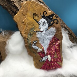 Op de afbeelding: Handgeschilderde rotskunst met een figuur in een rood-gouden jasje met patroon, witte kraag met ruches en zwarte hoed. De figuur houdt een klein glas vast. De rots heeft een goudkleurige achtergrond.
