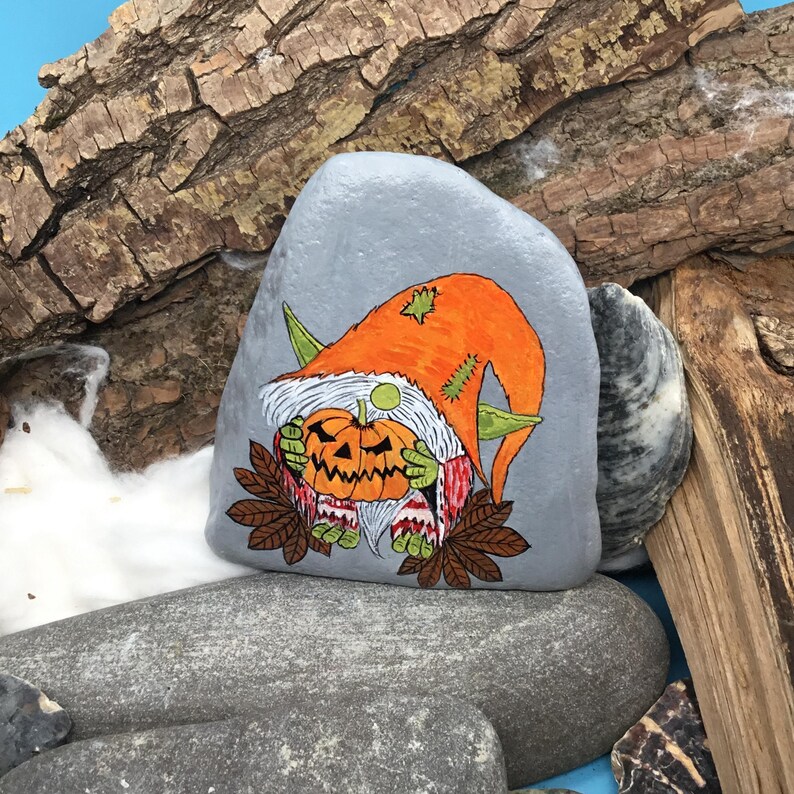 May include: A hand-painted, gray stone features a Halloween gnome holding a carved pumpkin. The gnome wears an orange hat and a red and white striped shirt. Brown leaves and green accents complete the design. The stone is displayed on a bed of rocks and wood.