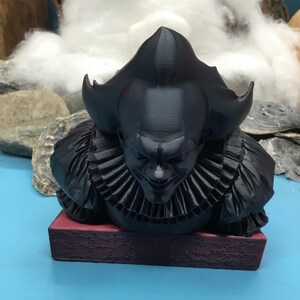 May include: A black 3D-printed bust of a menacing clown with a ruffled collar and a sinister expression. The bust is mounted on a rectangular, burgundy-colored base. The background features a blue surface and a backdrop of rocks and cotton.