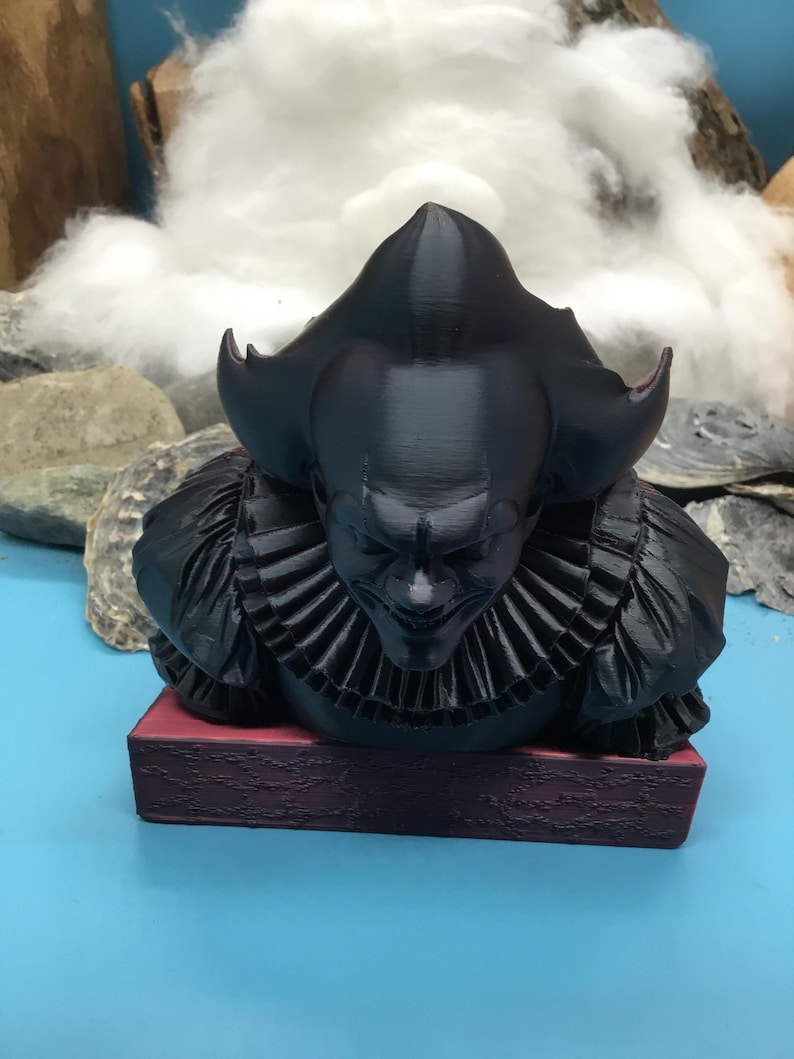 May include: A black 3D-printed bust of a menacing clown with a ruffled collar and a sinister expression. The bust is mounted on a rectangular, burgundy-colored base. The background features a blue surface and a backdrop of rocks and cotton.