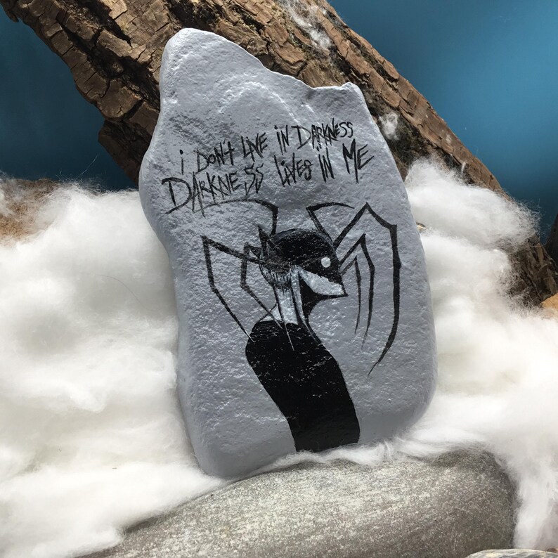 May include: A light gray painted rock featuring a black ink illustration of a stylized figure with long limbs and the hand-lettered text "I DON'T LIVE IN DARKNESS DARKNESS LIVES IN ME" written in a jagged, dark font across the upper portion of the stone.